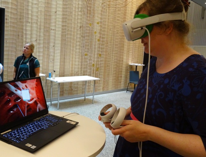 Sanna Innilällä on VR-lasit päässä ja ohjaimet käsissä. Kannettava tietokone pöydällä näyttää, että VR-laseista näkyy sydämen sisäpintoja.