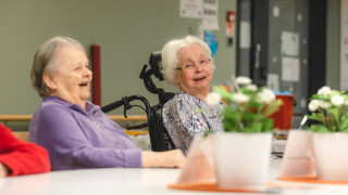 Kaksi ikääntynyttä naista nauravat pöydän ääressä.