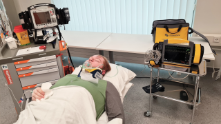 Simulaatioharjoituksessa potilas makaa sängyllä, hänellä on happimaski leuan päällä ja vieressä simulaatiossa käytettäviä laitteita.