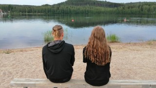 Kaksi henkilöä istuu puisella kapealla penkillä selin kameraan järven rannassa